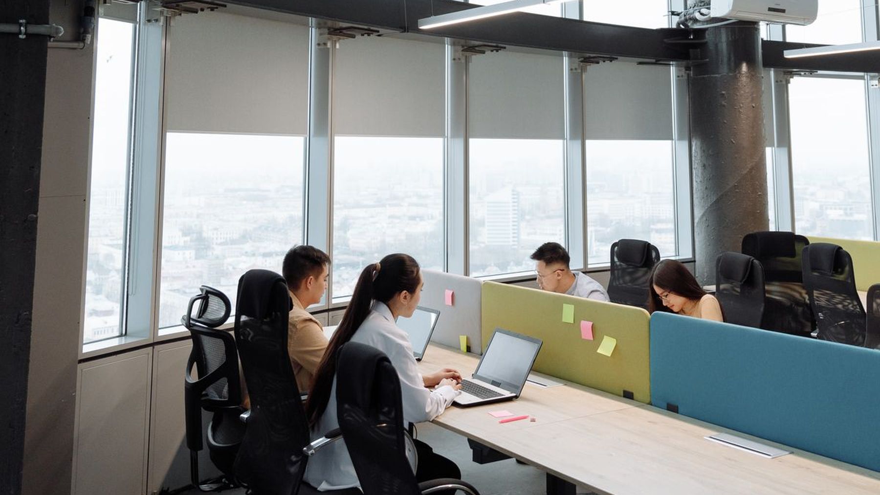 A high-rise office floor in daylight, people working at long desks beside a glass wall